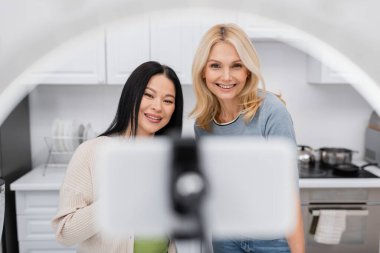 Cheerful interracial bloggers looking at blurred smartphone in ring lamp at home  clipart