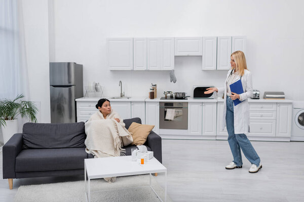 Doctor with clipboard pointing at sick asian patient at home 