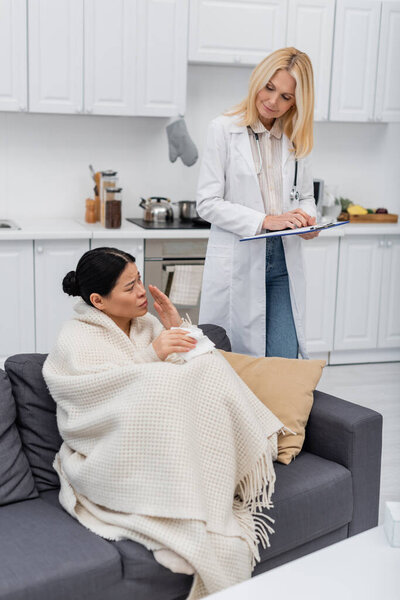 Ill asian woman in blanket talking to doctor with clipboard at home 