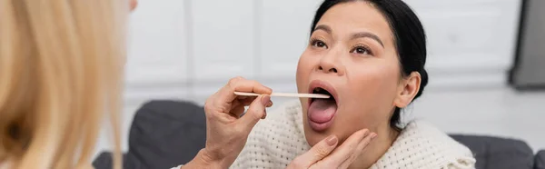 Blurred doctor holding tongue depressor near asian patient with open mouth at home, banner