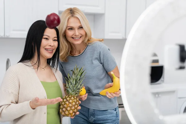 Asian blogger throwing apple near friend and smartphone in kitchen