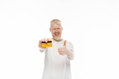 cheerful albino man in t-shirt holding credit card and showing thumb up isolated on white  clipart