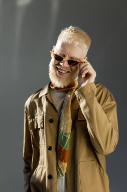 sunlight on face of cheerful albino man adjusting stylish sunglasses on grey  clipart