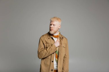 Sakallı albino adam şık gömlek ve ipek atkı giyerek gri renkte poz veriyor. 