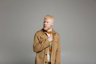 Sakallı albino adam şık gömlek ve ipek atkı giyerek gri renkte poz veriyor. 