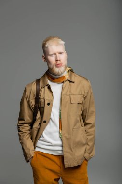 Şık gömlekli ve ipek atkılı sakallı albino adam elleri gri cebinde duruyor. 