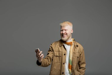 Sakallı ve mutlu albino ceketli adam gri üzerinde izole akıllı telefon kullanıyor.