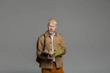 Sırt çantası olan sakallı albino öğrenci gri üzerinde not defteri tutuyor.