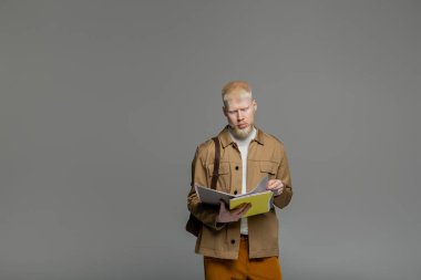 Sırt çantalı akıllı albino öğrenci gri üzerinde not defteri tutuyor.