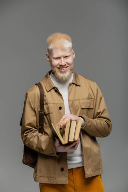 Sırt çantalı neşeli albino öğrenci gri üzerine kitap tutuyor.