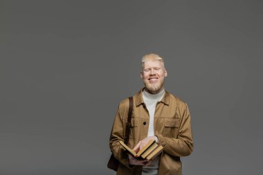Sırt çantalı mutlu albino öğrencisi gri üzerine kitap tutuyor.