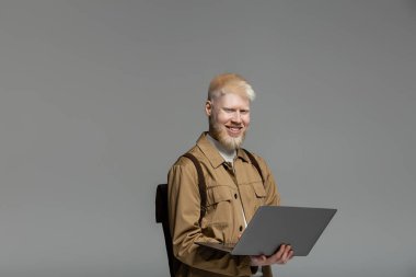 Sırt çantalı mutlu albino öğrencisi, internetten öğrenim görürken gri renkte izole edilmiş.