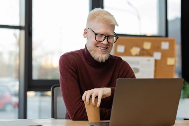 Gözlüklü neşeli albino işadamı dizüstü bilgisayara bakıyor ve ofisinde kağıt bardak tutuyor. 