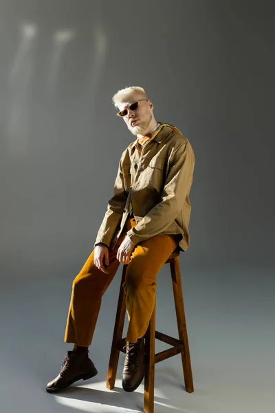 full length of stylish albino man in sunglasses and shirt jacket sitting on high chair on grey