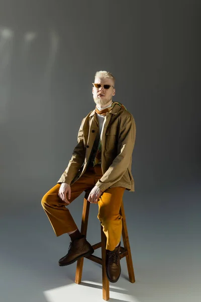 full length of bearded albino man in sunglasses and shirt jacket sitting on high chair on grey