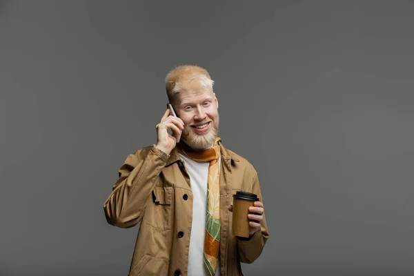 stock image happy albino man in shirt jacket holding paper cup and talking on smartphone isolated on grey