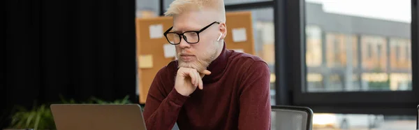 bearded albino businessman in earphones looking at laptop while working in office, banner