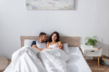 Man in pajama hugging brunette girlfriend in morning in bedroom 