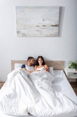 Young man hugging and looking at smiling girlfriend on bed 