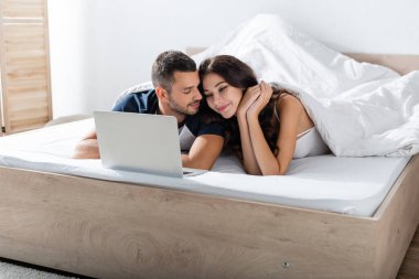 Bearded man looking at girlfriend near laptop on bed at home 