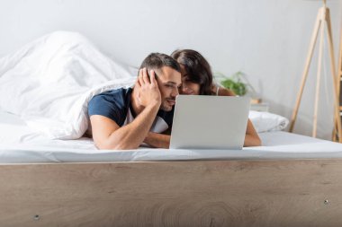 Young man looking at laptop near brunette girlfriend on bed 
