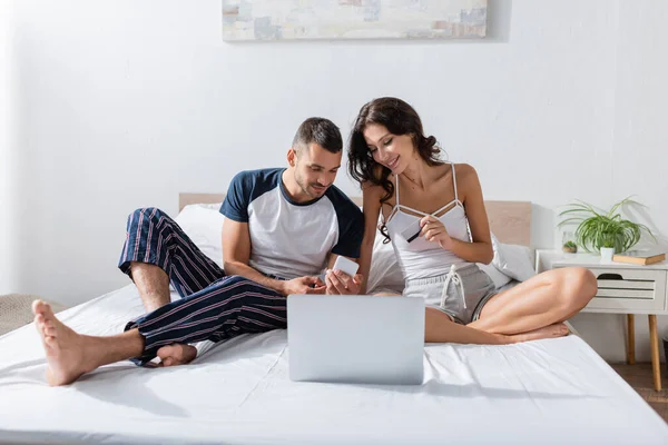Smiling couple with credit card using devices on bed at home