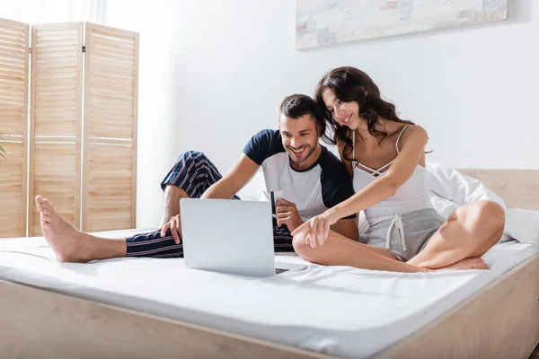 Smiling woman pointing at laptop near boyfriend with credit card on bed