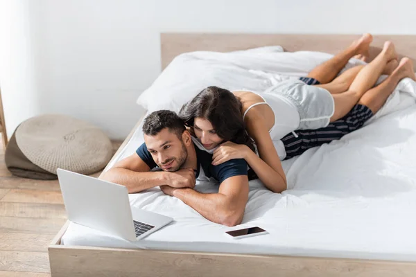 Couple looking at laptop near smartphone on bed at home