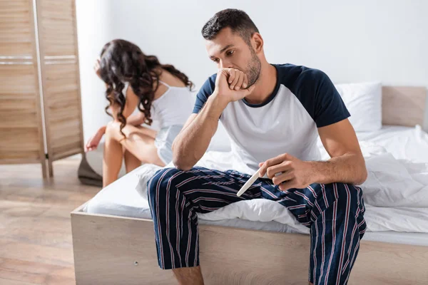 worried man in pajama holding pregnancy test near blurred girlfriend at home
