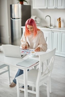 Pembe saçlı, sıradan giyinmiş, laptopla çalışan ve resimlerle uğraşan neşeli genç bir kadın.