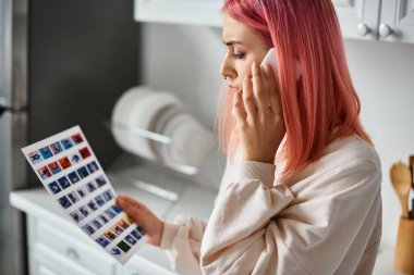 good looking pink haired woman in everyday attire talking by phone and looking at paper with images clipart