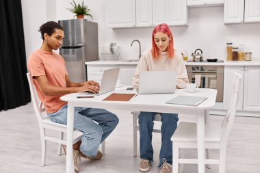 Güzel görünümlü, çok kültürlü, ev elbiseli çift dizüstü bilgisayarlarını evde uzaktan çalışmak için kullanıyorlar.