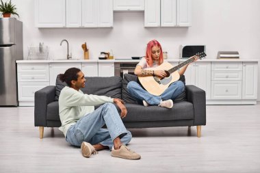 Neşeli Afro-Amerikalı ev elbiseli adam pembe saçlı kız arkadaşının gitar çalışına sevgiyle bakıyor.