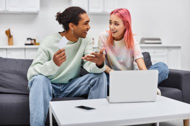 joyful interracial couple in homewear holding credit card and smiling at each other near laptop clipart