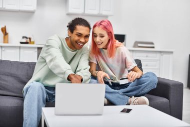 Kredi kartı tutan ve evdeki oturma odasında dizüstü bilgisayara bakan neşeli çift.