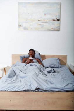 Genç bir Afro-Amerikalı, sabahları cep telefonu ekranına takılı olarak yatakta yatıyor..