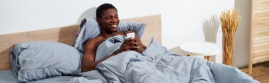 A young African American man lying in bed, fully engaged with his cell phone in the morning. clipart