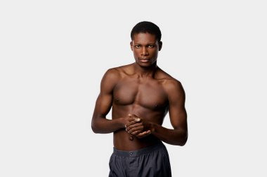 Afro-Amerikalı bir adam, stüdyo ortamında beyaz bir zemine karşı gömleksiz duruyor..