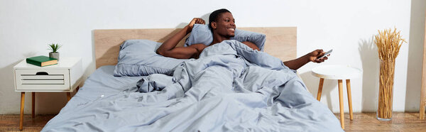 A young African American man lays in bed, holding a cell phone in his hand, connecting with the digital world upon waking.