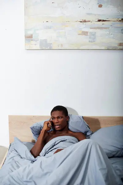 stock image A young African American man lays in bed, talking on a cell phone in the morning light.