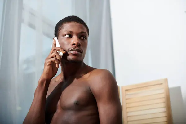 stock image A young African American man is shirtless, deeply engrossed in conversation on his cell phone.