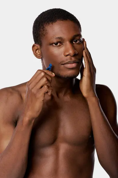 stock image African American man without shirt shaving with razor in studio on white background.