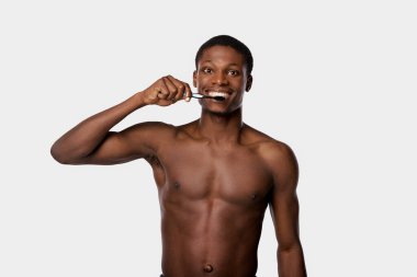 Shirtless African American man in studio brushing his teeth with a toothbrush against a white background. clipart