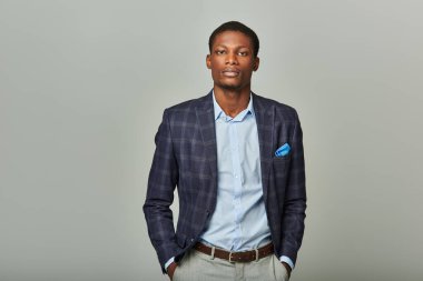Yakışıklı Afrikalı Amerikalı iş adamı, elleri ceplerinde gri arka planda damalı ceket giyerek güvenle duruyor..