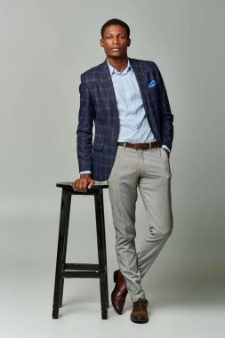 Resmi giyinmiş yakışıklı Afro-Amerikan adam gri bir arka planda bir tabureye yakın duruyor..