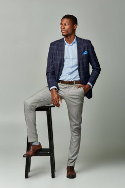 Ekoseli ceketli yakışıklı Afro-Amerikan adam gri arka planda resmi giyinmiş olarak taburede oturuyor..