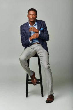 Siyah ceketli yakışıklı Afro-Amerikan erkek, gri arka planda taburede oturuyor..