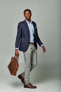 An elegant African American businessman in a checkered blazer holding a brown bag on a grey background. clipart