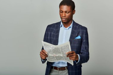 Kareli ceketli yakışıklı Afro-Amerikan işadamı gri arka planda gazeteye gömüldü..