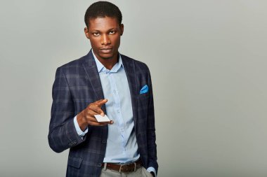 Takım elbiseli yakışıklı Afro-Amerikan işadamı gri arka planda iş kartı tutuyor..